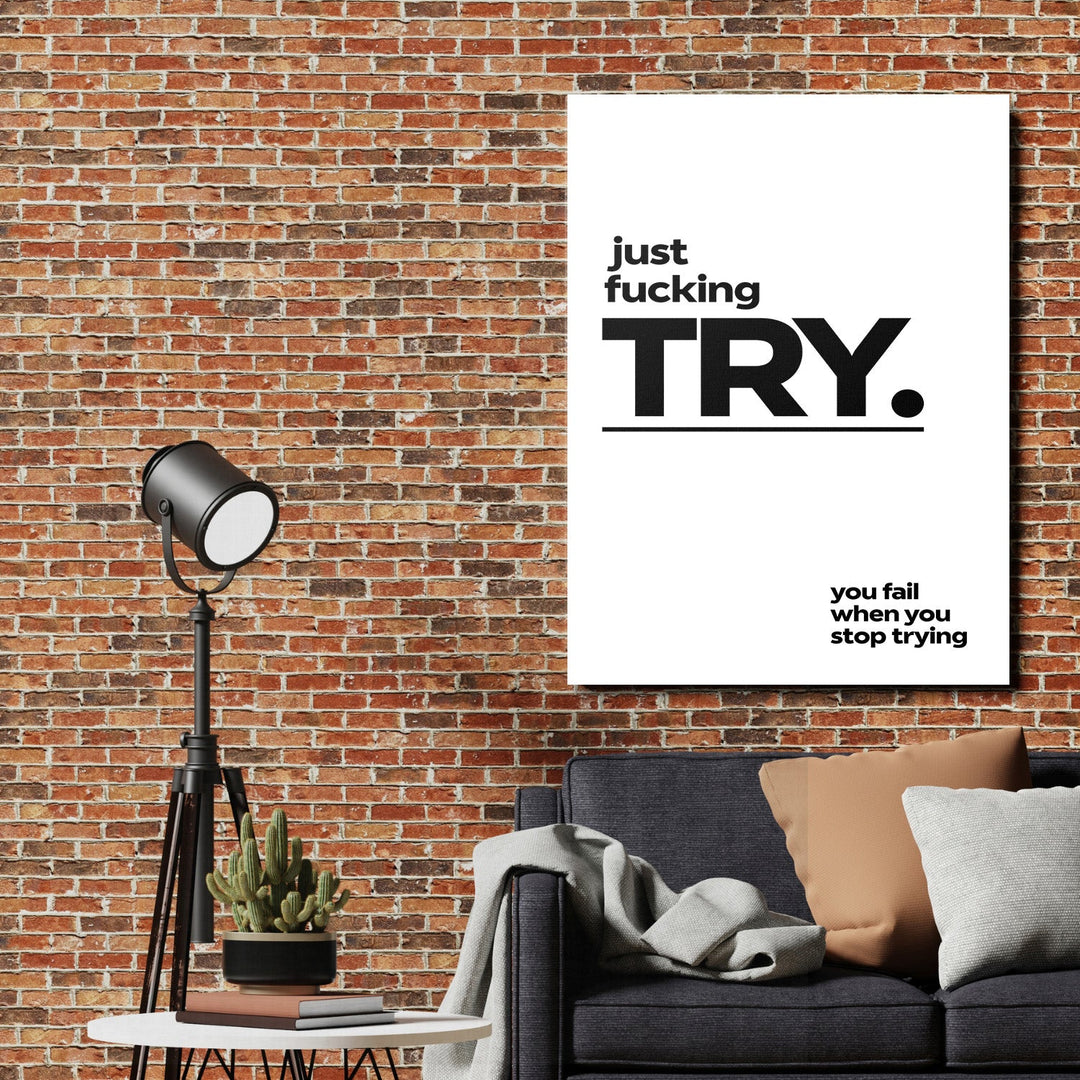 Motivational canvas print “just fucking TRY” on a brick wall in a living room above a sofa and floor lamp