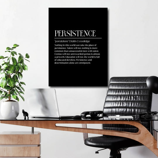 Motivational wall art with a black-and-white quote about persistence above a desk with a flower and open laptop